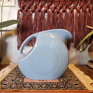 May include: A light blue ceramic pitcher with a large handle and a spout. The pitcher has a rounded body with concentric circle detailing. It sits on a small rug with a geometric pattern and fringe.