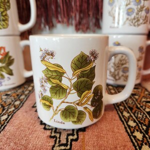 Op de afbeelding: Witte keramische mok met een groen-wit bloemenontwerp en het woord "Linde" gedrukt op de bodem. De mok staat op een kleurrijk tapijt.