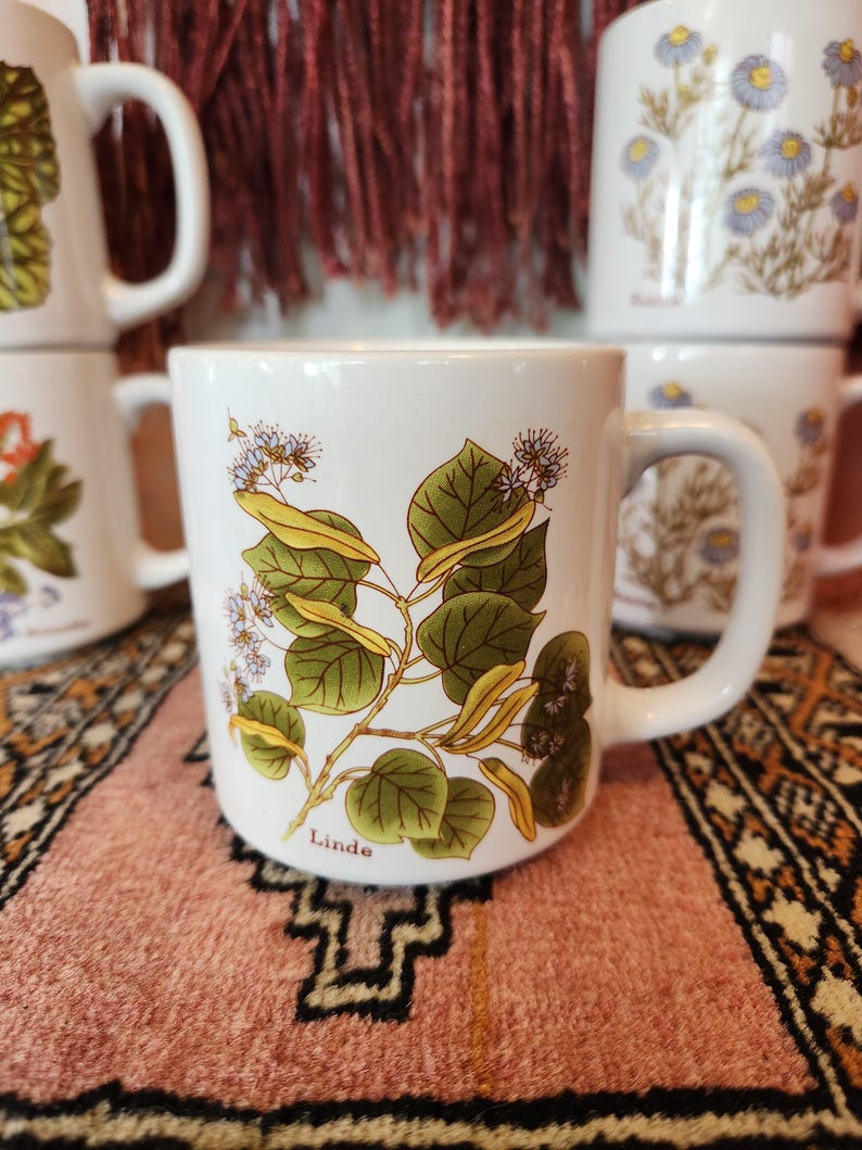 Op de afbeelding: Witte keramische mok met een groen-wit bloemenontwerp en het woord "Linde" gedrukt op de bodem. De mok staat op een kleurrijk tapijt.