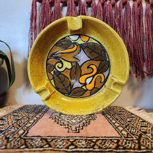May include: Yellow ceramic ashtray with a central design of brown leaves and orange and white abstract shapes. The ashtray has four built-in rests for cigarettes and is displayed on a patterned rug.