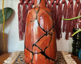 May include: A tall, orange ceramic vase with a glossy finish. The vase features a unique design of black lines that resemble cracks. The vase is displayed on a patterned rug, with a woven wall hanging in the background.