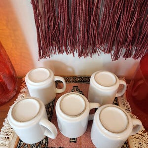 Op de afbeelding: Zes witte keramische mokken met handvatten, gerangschikt op een roze en bruin gepatroneerd kleed met franjes. De mokken zijn allemaal even groot en even gevormd.