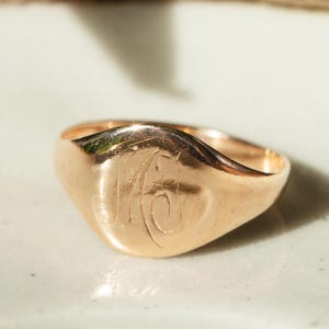 May include: A polished rose gold signet ring featuring a smooth, rounded face with an engraved monogram consisting of the letters M and J intertwined in a cursive script. The metal surface exhibits a warm, metallic lustre and subtle reflections.