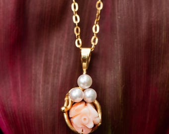 May include: A gold necklace with a pendant. The pendant features a carved coral flower and three white pearls. The necklace has a delicate chain and is set against a dark red background.