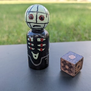 May include: A hand-painted wooden doll with a black and white skeleton design and a green face. The doll is standing next to a small wooden box with a black and white design.