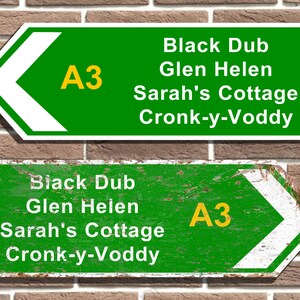 May include: Two green road signs with white arrows pointing left. The signs have white text on a green background and yellow text that reads "A3". The top sign is newer and the bottom sign is weathered and rusty. The text on the signs reads: "Black Dub, Glen Helen, Sarah's Cottage, Cronk-y-Voddy."
