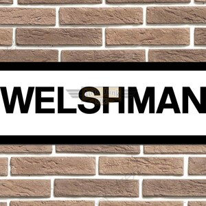May include: A rectangular sign with the word "WELSHMAN" in bold, black letters on a white background, framed by a black border. The sign is mounted on a brick wall with varying shades of brown.