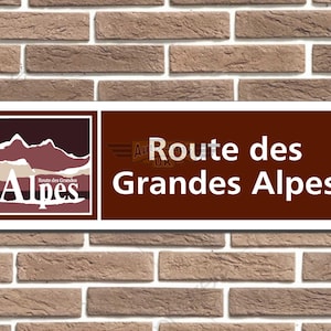 Könnte beinhalten: Rechteckiges Schild mit dem Text "Route des Grandes Alpes" in weißen Buchstaben auf braunem Hintergrund. Das Schild zeigt auch ein Logo mit Bergsilhouetten und dem Wort "Alpes". Das Schild ist an einer Ziegelmauer befestigt.