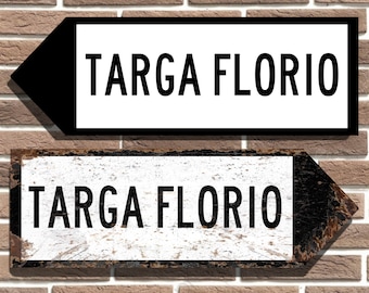 Puede incluir: Dos señales con flechas negras que apuntan en direcciones opuestas. Ambas señales son blancas con texto negro que dice "TARGA FLORIO". La señal superior es más nueva y tiene un borde negro. La señal inferior es más antigua y tiene un marco de madera desgastado.
