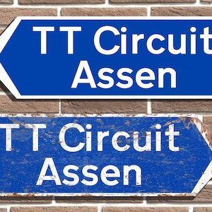 May include: Two blue signs with white text, one new and one worn, both pointing to the left and reading "TT Circuit Assen".