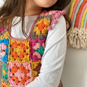 May include: A colourful crocheted vest with a square pattern in shades of pink, orange, yellow, green, and brown. The vest is sleeveless and has a rounded neckline.
