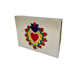Op de afbeelding: Een zilverkleurige rechthoekige doos met een decoratief hartontwerp. Het hart is rood, omgeven door een gele rand en versierd met blauwe, rode en groene accenten. De doos heeft een scharnierend deksel.