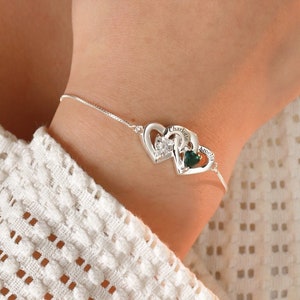 May include: A silver bracelet with two interlocking heart charms. One heart features a small green gemstone, while the other has a small clear gemstone. The bracelet is worn on a person's wrist.
