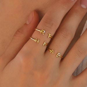 Puede incluir: Tres anillos de oro con las letras R, T, H, F, A y E en ellos. Los anillos se llevan en el dedo de una persona.