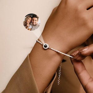 以下が含まれることがあります： 中央に小さな丸い黒い石が付いたシルバーブレスレット。ブレスレットは繊細なチェーンデザインで、小さなエクステンションチェーンが付いています。小さな円形の画像が腕に投影されています。