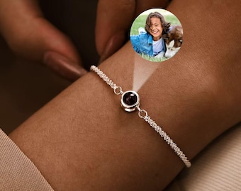 May include: A silver bracelet with a small, round, dark red gemstone at the center. The bracelet's chain has a textured, twisted design. The image includes a small circular photo of a person.