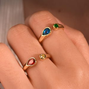 Puede incluir: Anillos dorados con piedras preciosas de colores. Los anillos presentan piedras azules y rojas en forma de lágrima, y una piedra amarilla redonda. También se ve una piedra verde. Los anillos son abiertos.