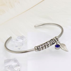 Puede incluir: Un brazalete de plata con un dije que tiene una piedra preciosa azul y un diseño de hoja. El brazalete tiene una serie de cuentas con nombres grabados en ellas: Ayla, Eder, Elme, Yirke, Iris, Tara.