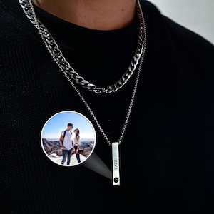 May include: A silver-toned layered necklace featuring a thick curb chain and a thinner chain with a vertical bar pendant. The bar is engraved with the word FAITH and contains a small circular projection lens displaying a photograph of a couple.