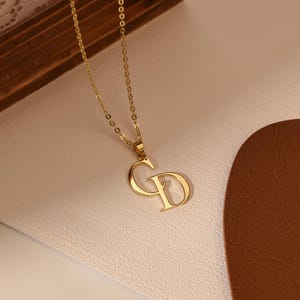 Puede incluir: Un collar dorado con un colgante que muestra las letras entrelazadas "CD". La cadena es fina y el colgante está en una fuente estilizada. La joya se presenta sobre una superficie de color claro. Es un accesorio de moda.