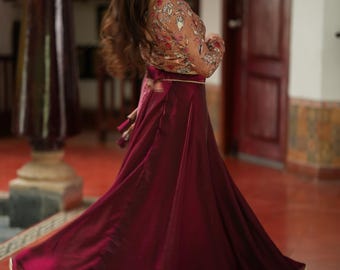May include: A flowing, burgundy skirt with a gold trim is paired with a sheer, long-sleeved top adorned with floral embroidery. The outfit is worn by a person in a room with red tile flooring.