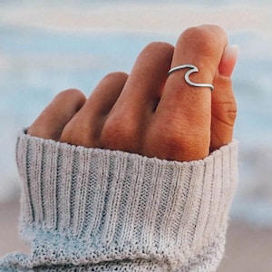 Könnte beinhalten: Ein silberner Wellenring wird an einem Finger getragen. Das Design des Rings ahmt eine sich kräuselnde Meereswelle nach. Die Hand wird von einem hellgrauen Strickpullover bedeckt, mit einem verschwommenen Ozeanhintergrund.
