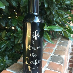 Puede incluir: Botella de vino de acero inoxidable negro con una inscripci&oacute;n plateada que dice "Life is better at the Lake" con una silueta de una persona esquiando en el agua.