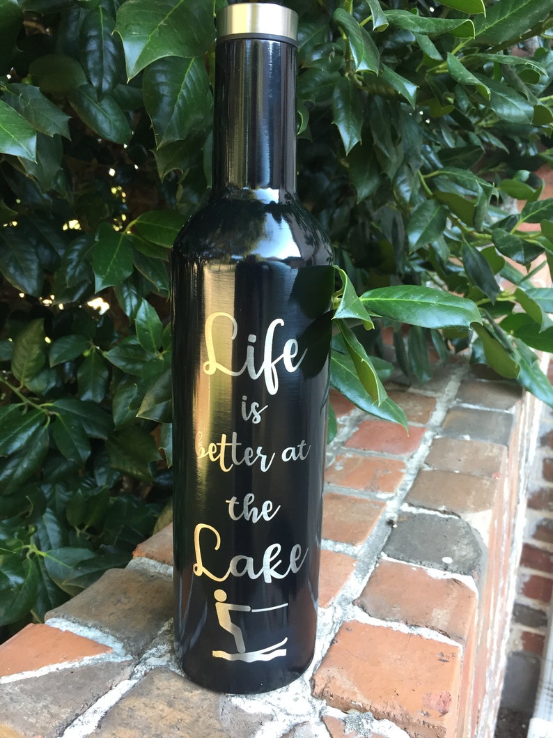 Puede incluir: Botella de vino de acero inoxidable negro con una inscripci&oacute;n plateada que dice "Life is better at the Lake" con una silueta de una persona esquiando en el agua.