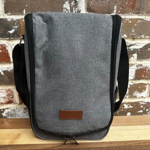 Puede incluir: Una bolsa de vino aislada de color gris con un parche de cuero marr&oacute;n y una cremallera negra. La bolsa tiene una correa de hombro negra ajustable.