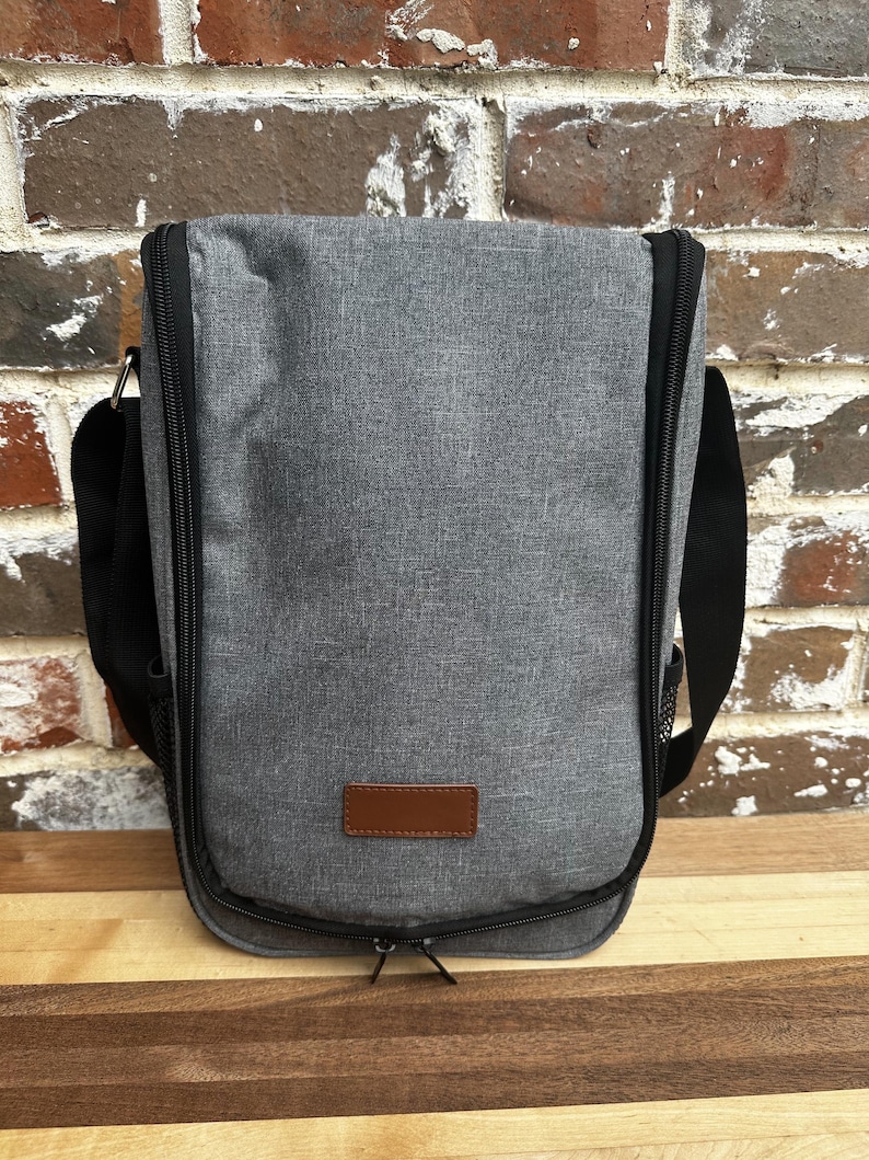 Puede incluir: Una bolsa de vino aislada de color gris con un parche de cuero marr&oacute;n y una cremallera negra. La bolsa tiene una correa de hombro negra ajustable.