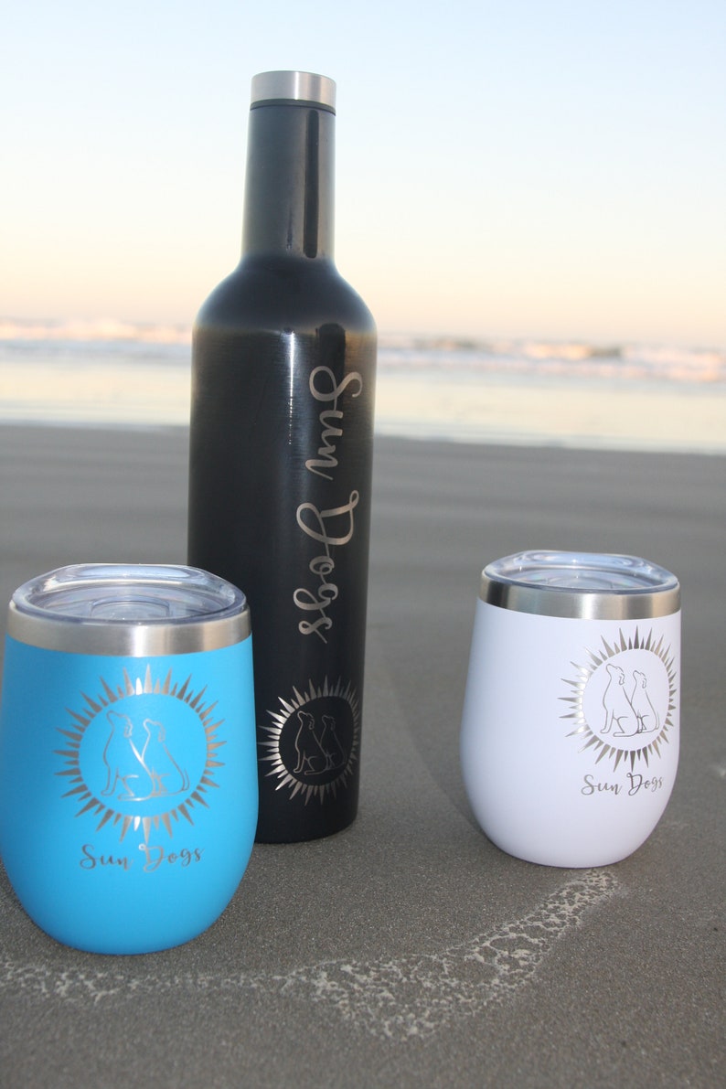 Puede incluir: Dos vasos de acero inoxidable con un dise&ntilde;o de sol y dos perros, uno azul y otro blanco, y una botella de acero inoxidable negra con el texto "Sun Dogs" en ella.
