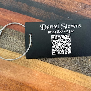 May include: Black metal luggage tag with a silver cable loop. The tag has the name "Darrel Stevens" and a phone number (214) 807-5412 engraved on it. A black and white QR code is also on the tag.