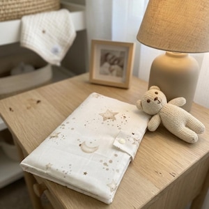 Peut inclure: Un matelas à langer en tissu rectangulaire et plié, doté d'une base crème avec un motif d'étoiles, de lunes et de points beiges. Le matelas est fixé par un bouton-pression blanc et est accompagné d'un petit ours en peluche tricoté beige.