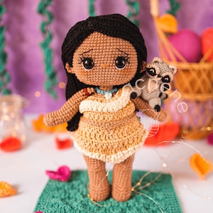 Puede incluir: Muñeca de ganchillo hecha a mano con cabello negro y vestido beige, sosteniendo un pequeño mapache gris. La muñeca está sobre un cuadrado verde, rodeada de hojas de ganchillo coloridas y una cesta en el fondo. La muñeca mide unos 20 cm.