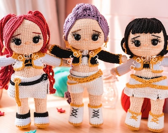 Puede incluir: Tres muñecas tejidas a ganchillo a mano con cabello rojo, morado y negro. Cada muñeca viste un conjunto de dos piezas blanco y dorado con detalles negros, zapatos blancos y pendientes de aro dorados. Las muñecas tienen ojos grandes y oscuros, rasgos bordados y brazos extendidos.