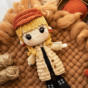 Puede incluir: Una muñeca de crochet que lleva un abrigo beige, un gorro naranja y botas negras. La muñeca tiene el pelo rubio y grandes ojos negros. La muñeca está sentada sobre una superficie peluda marrón.