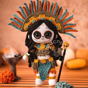 May include: Crocheted Day of the Dead doll with a colorful headdress, skull face, and long black hair. The doll is holding a staff with a skull on top. The doll is standing on a wooden surface with orange and green pom-poms.