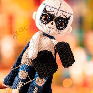 May include: A white crocheted doll with a black and blue face mask, wearing a blue and gray cape and black gloves. The doll has a skull-like face with black eyes and a stitched mouth.
