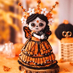 Puede incluir: Muñeca de ganchillo hecha a mano al estilo de una figura del Día de Muertos. La muñeca presenta un vestido negro y naranja, una corona floral y una cara pintada. Se exhibe sobre una rodaja de madera.