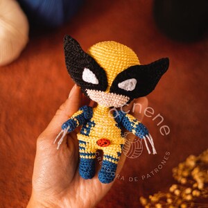 Puede incluir: Un juguete de amigurumi tejido a crochet de Wolverine de los X-Men. El juguete es amarillo con detalles azules y tiene una máscara negra con ojos blancos. El juguete está siendo sostenido en la mano de una persona.
