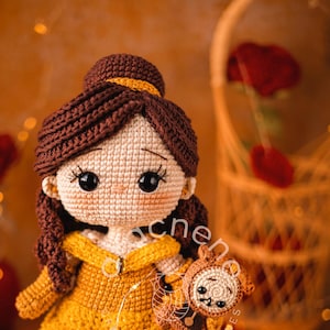 Puede incluir: Una muñeca de ganchillo de Bella de La Bella y la Bestia, con un vestido amarillo y sosteniendo un peluche de ganchillo de la Bestia. La muñeca tiene el pelo castaño y grandes ojos marrones.