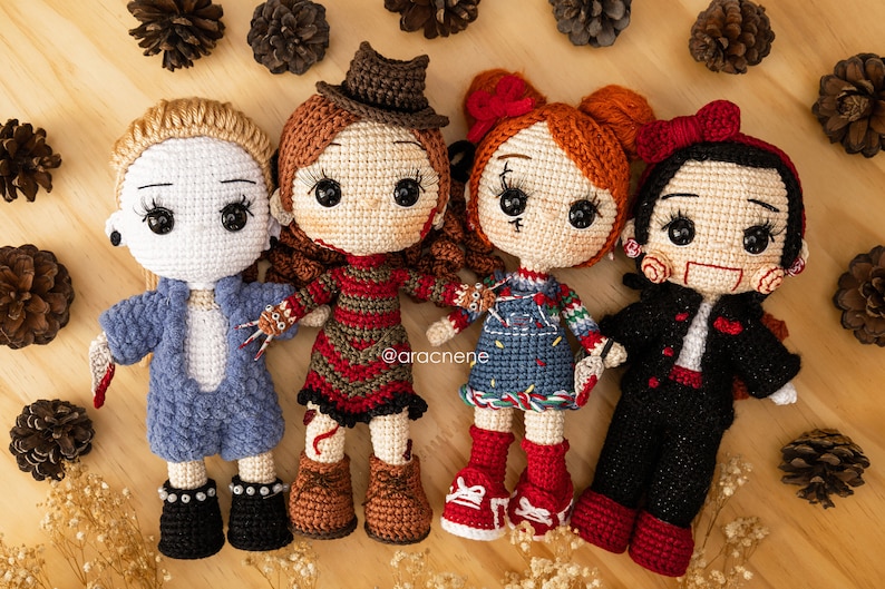 K&ouml;nnte beinhalten: Vier geh&auml;kelte Puppen, die als Horrorfilmfiguren verkleidet sind. Die Puppen stehen auf einer Holzoberfl&auml;che. Die Puppen tragen rote, blaue, schwarze und braune Kleidung. Die Puppen haben schwarze Augen und rote Lippen. Die Puppen halten Requisiten aus den Filmen, als die sie verkleidet sind. Die Puppen l&auml;cheln alle.
