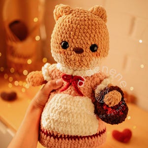 May include: A crocheted stuffed animal in the shape of a fox wearing a pink dress with a white apron and a red bow. The fox is holding a brown crocheted heart in its paw. The fox is surrounded by red hearts.