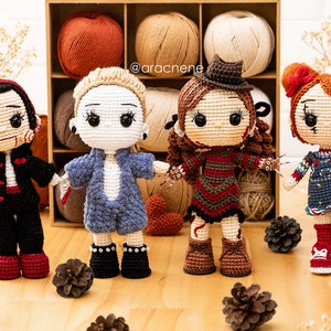 K&ouml;nnte beinhalten: Vier geh&auml;kelte Puppen, die als Horrorfilmfiguren verkleidet sind. Die Puppen stehen auf einer Holzoberfl&auml;che mit Tannenzapfen. Die Puppen tragen verschiedene farbige Outfits und haben verschiedene Frisuren. Die Puppen l&auml;cheln alle. Der Text '@aracnene' ist im Hintergrund sichtbar.