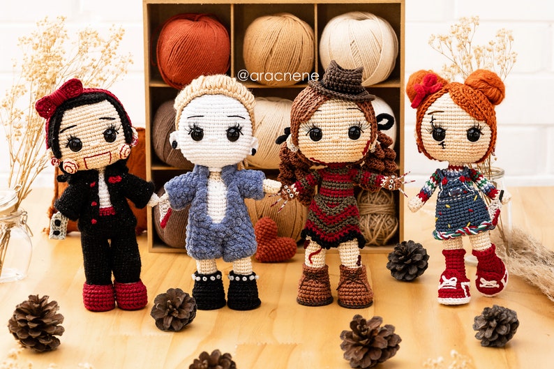 K&ouml;nnte beinhalten: Vier geh&auml;kelte Puppen, die als Horrorfilmfiguren verkleidet sind. Die Puppen stehen auf einer Holzoberfl&auml;che mit Tannenzapfen. Die Puppen tragen verschiedene farbige Outfits und haben verschiedene Frisuren. Die Puppen l&auml;cheln alle. Der Text '@aracnene' ist im Hintergrund sichtbar.