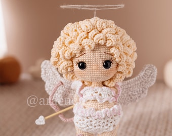 May include: A crocheted doll featuring curly blonde hair, a white halo, and grey wings. The doll wears a white ruffled outfit with pink accents, holds a small wooden bow with a heart-tipped arrow, and has small pink heart details on its legs.