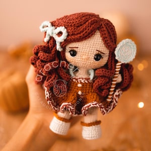 Puede incluir: Una muñeca de crochet con pelo marrón, un vestido marrón y blanco y botas blancas. La muñeca sostiene una piruleta azul y blanca.