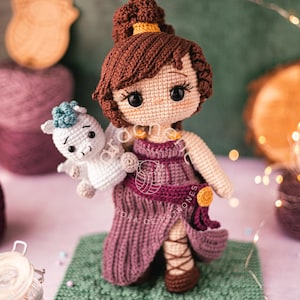 May include: A handmade crochet doll with brown hair and a purple dress, holding a white crochet Pegasus. The doll is standing on a green square, with yarn and decorative lights in the background. The doll's hair is styled in a bun.