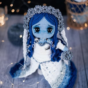 May include: A handmade crochet doll in the style of a bride, featuring blue skin, dark eyes, and blue hair. The doll wears a white and blue dress with a floral headpiece. The doll is holding a small bouquet. The text "arochene" is visible.