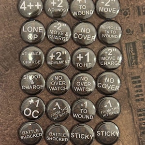 Könnte beinhalten: Eine Sammlung von einundzwanzig runden Spielmarken mit weißen Texten. Die Marken zeigen Sätze wie "+2 Wounds", "-2 Move & Charge" und "Sticky". Die Marken sind dunkel mit weißer Schrift.